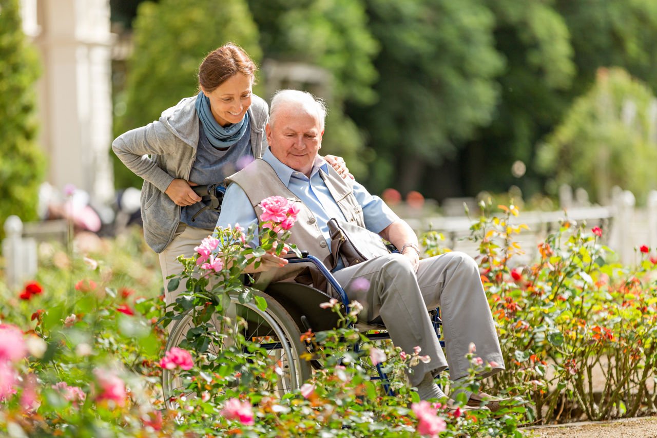 Kvinne trillar ein eldre mann i rullestol gjennom ein hage. Mannen ser og tar på rosene.