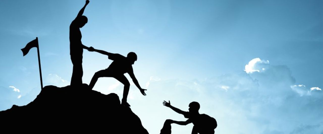 Bildet viser silhuetten av ein person på ein topp, som hjelper ein annan person opp til seg